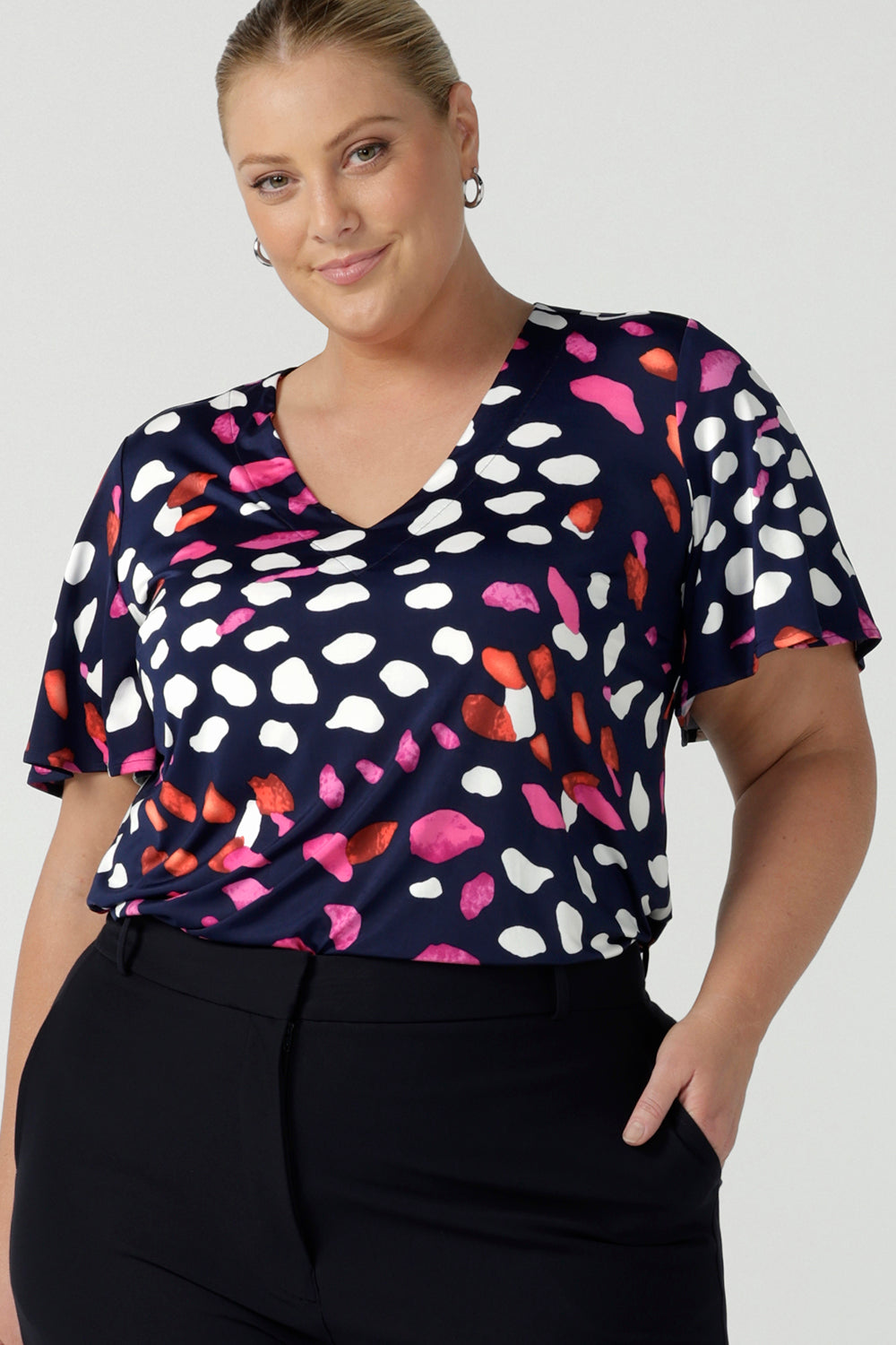 A plus size, size 18 woman wears a abstract print jersey, V-neck top with flutter sleeves. A good top for summer casual wear, or style tucked as a workwear top. Shop made in Australia tops in petite to plus sizes online at Australian fashion brand, Leina & Fleur.