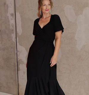 Showing a good black dress for women, an over 40, size 10 woman wears a black maxi length wrap dress with flutter sleeves. Made in Australia by women's clothing label, Leina & Fleur, this black cocktail dress is a great dress for curvy, mid size and petite women's evening and event wear.