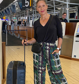 Showing great travel outfits for long weekend getaways, an over 50 woman is showing her airport style in wide leg printed pants and a 3/4 sleeve,V-neck jersey top in charcoal. All her travel clothing is made in Australia by Australian and New Zealand women's clothing brand, Leina and Fleur and available to shop online in sizes 8 to 24.