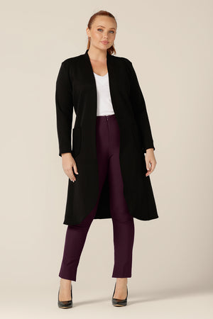 A curvy size 12 woman wears a softly tailored black trenchcoat in modal fabric. Worn with mulberry slim leg pants and a white bamboo jersey top, this is a great coat for winter layering and travel.