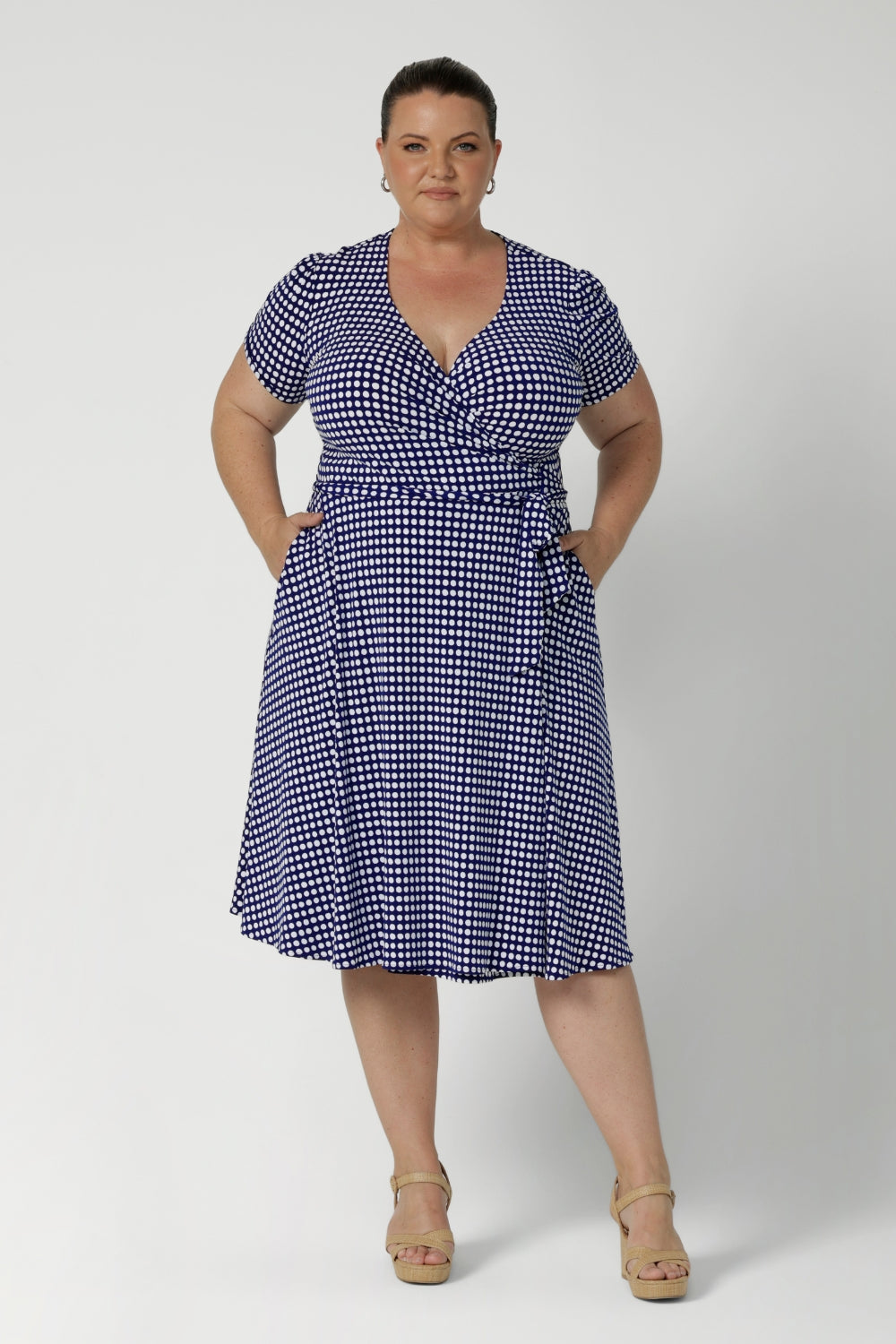 Plus size woman wears polka dotted wrap dress. Knee length dress is petite height and plus size friendly, available in sizes across 8-24.  Short sleeve dress features navy polka dot print, wrap design and pockets. Made in Australia from easy care, wrinkle resistant, dry touch jersey fabric. Crafted by woman's fashion brand Leina & Fleur.