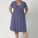 Plus size woman wears polka dotted wrap dress. Knee length dress is petite height and plus size friendly, available in sizes across 8-24.  Short sleeve dress features navy polka dot print, wrap design and pockets. Made in Australia from easy care, wrinkle resistant, dry touch jersey fabric. Crafted by woman's fashion brand Leina & Fleur.