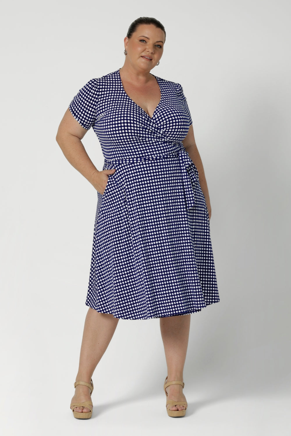 Knee length dress is petite height and plus size friendly, available in sizes across 8-24. Size 18 model wears short sleeve, navy polka dot printed dress with nude heels. Wrap dress is made from wrinkle resistant fabric that is easy care. Made in Australia by woman's clothing brand Leina & Fleur.