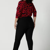 Back view of a size 18 woman wears the Mason top in Bold Poppy . A long sleeve jersey top for women with red floral on a black base. Comfortable work top for women size 8 - 24. Soft jersey fabric and made in Australia for women. Styled back with Black Brooklyn slim fit pants and black heels.