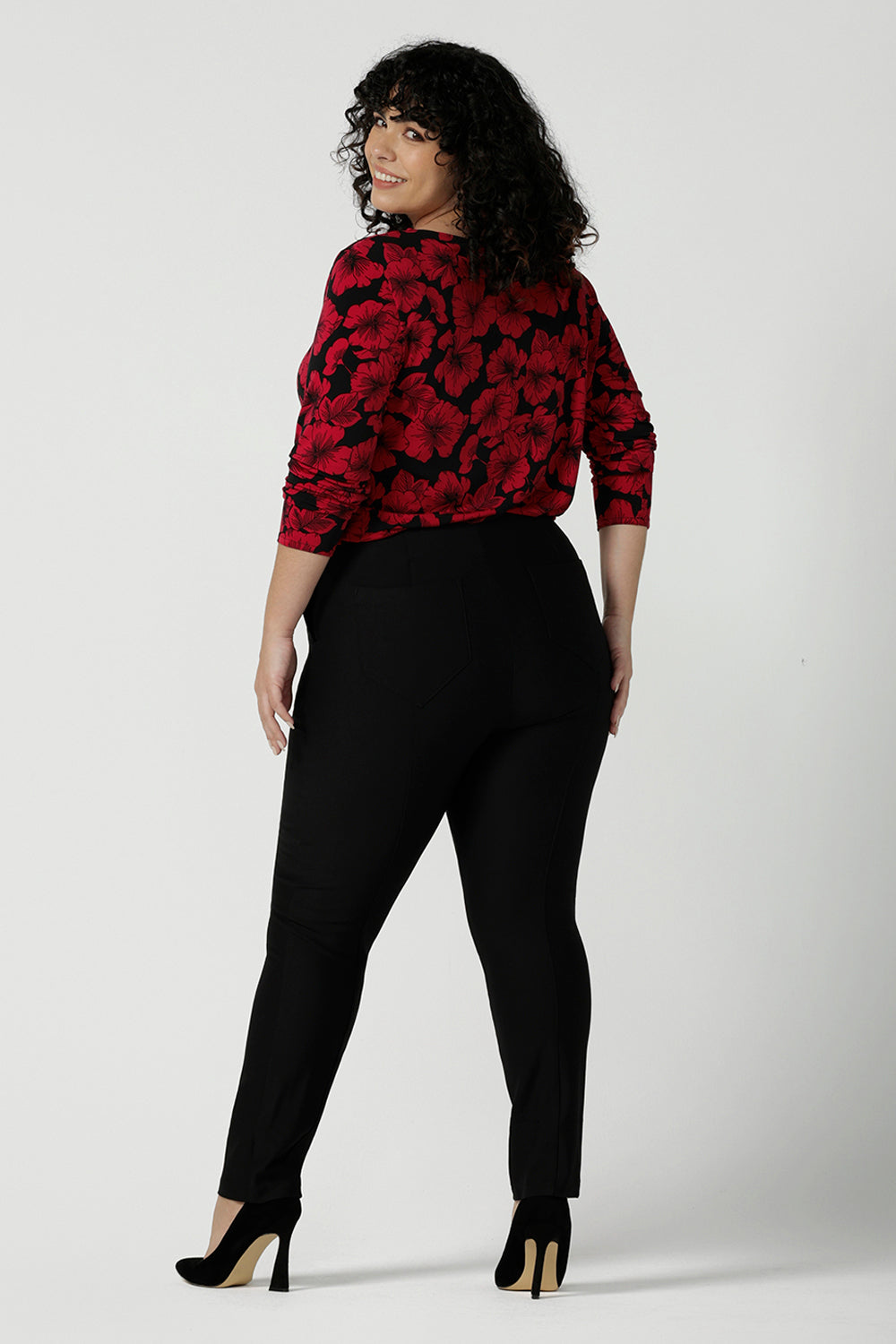 Back view of a size 18 woman wears the Mason top in Bold Poppy . A long sleeve jersey top for women with red floral on a black base. Comfortable work top for women size 8 - 24. Soft jersey fabric and made in Australia for women. Styled back with Black Brooklyn slim fit pants and black heels.