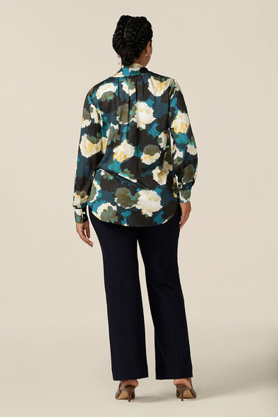 Back view of a luxe pull-on shirt in abstract floral print, the Matisse Shirt in Pom Pom has a V-neckline with pussy bow neck ties and long, bishop sleeves that blouson over fitted cuffs. Worn with flared leg, tailored navy pants to show this workwear blouse's shirttail hemline.