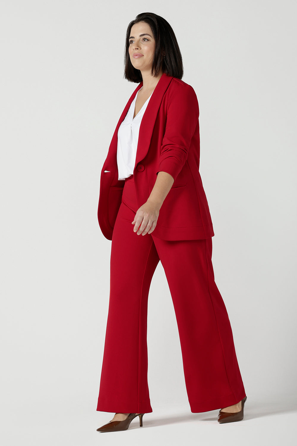 A size 10 woman wears the Merit Blazer in Flame, a textured scuba crepe blazer with button front. Curved hemline and front pockets. Made in Australia for women size 8 - 24. Stylish corporate comfortable. Styled back with matching red wide leg pants.