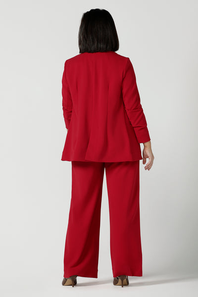 Back view of a size 10 woman wears the Merit Blazer in Flame, a textured scuba crepe blazer with button front. Curved hemline and front pockets. Made in Australia for women size 8 - 24. Stylish corporate comfortable. Styled back with matching red wide leg pants.