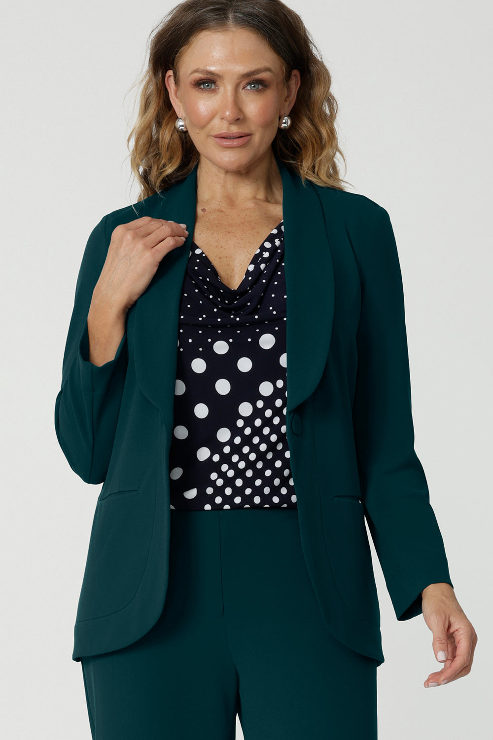 Showing a good work blazer for over 50s women, a size 12 woman wears a longline, green blazer with pockets. Worn with a navy and white spot print top and wide leg green workwear pants, this soft suiting is perfect for work and corporate wear. Made in Australia by Australian and New Zealand women's fashion label, Leina & Fleur, shop workwear blazers in plus size to petite height in their online boutique.