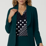 Showing a good work blazer for over 50s women, a size 12 woman wears a longline, green blazer with pockets. Worn with a navy and white spot print top and wide leg green workwear pants, this soft suiting is perfect for work and corporate wear. Made in Australia by Australian and New Zealand women's fashion label, Leina & Fleur, shop workwear blazers in plus size to petite height in their online boutique.