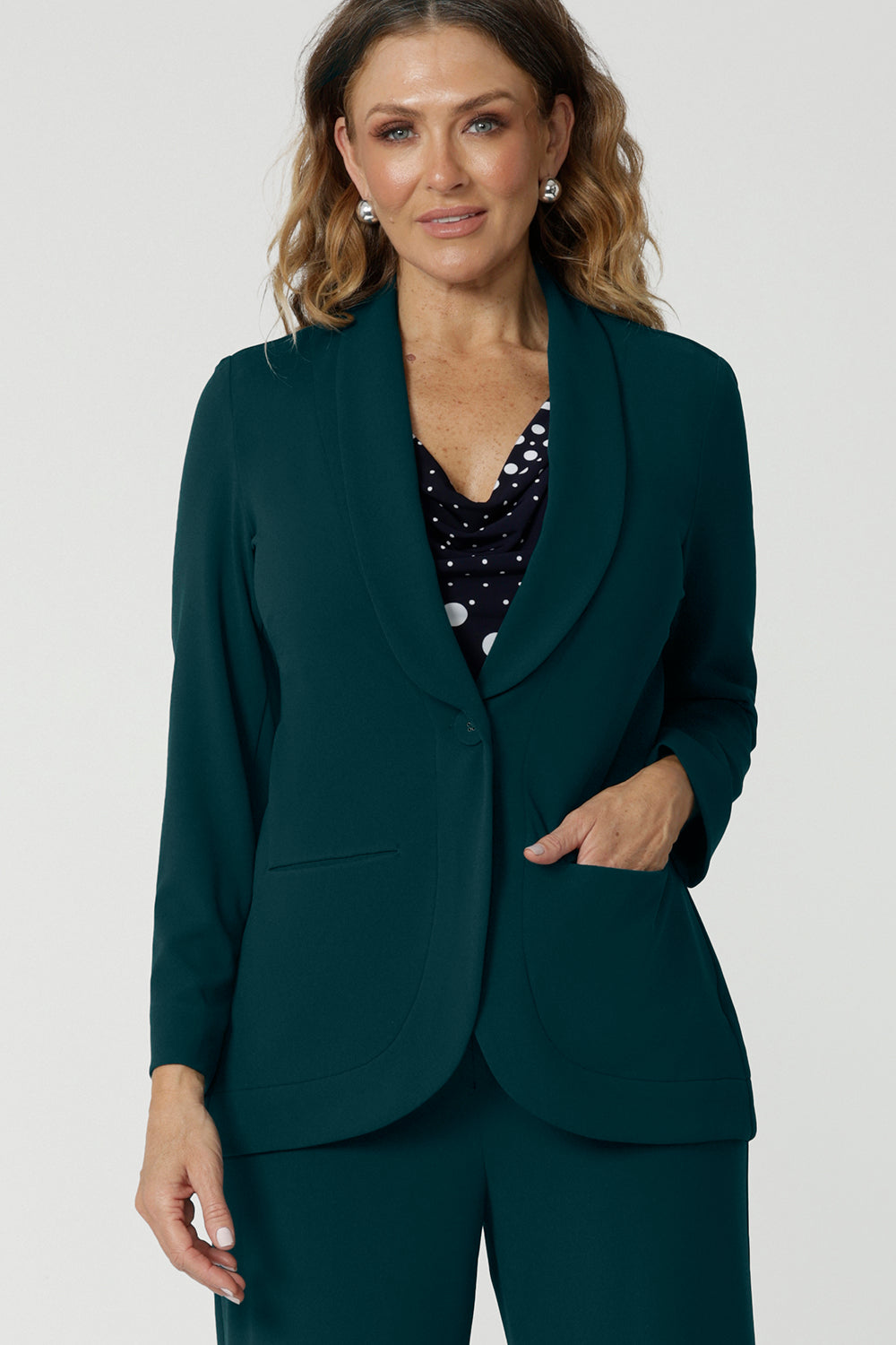 Showing a good work blazer for over 50s women, a size 12 woman wears a longline, green blazer with pockets. Worn with a navy and white spot print top and wide leg green workwear pants, this soft suiting is perfect for work and corporate wear. Made in Australia by Australian and New Zealand women's clothing brand, Leina & Fleur, shop work blazers in plus size to petite height in their online boutique.