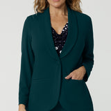 Showing a good work blazer for over 50s women, a size 12 woman wears a longline, green blazer with pockets. Worn with a navy and white spot print top and wide leg green workwear pants, this soft suiting is perfect for work and corporate wear. Made in Australia by Australian and New Zealand women's clothing brand, Leina & Fleur, shop work blazers in plus size to petite height in their online boutique.