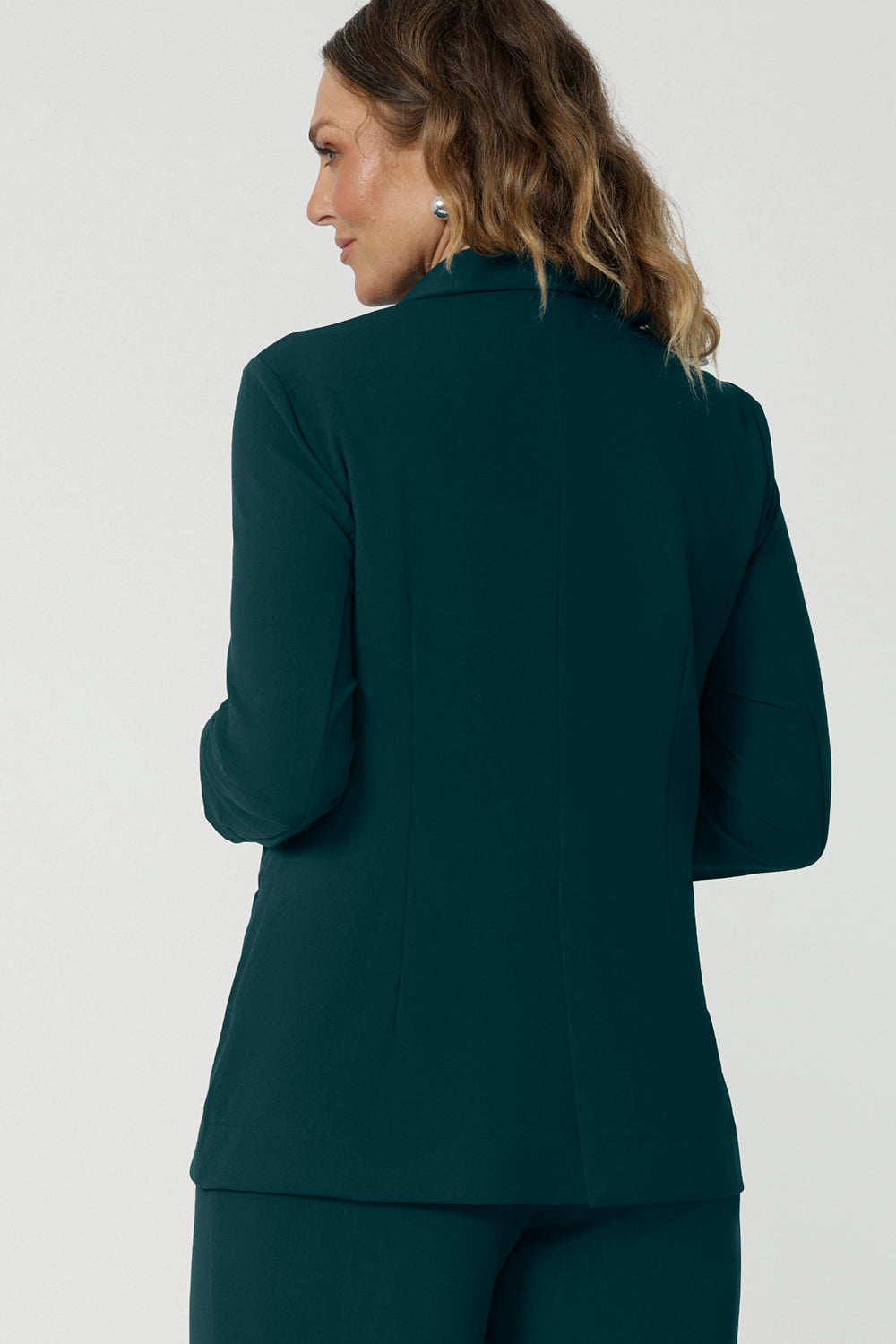 Back view of a good work blazer for over 50s women . A size 12 woman wears a longline, green blazer with pockets. Worn with wide leg green workwear pants, this stretch jacket  is perfect for work and corporate wear. Made in Australia by Australian and New Zealand women's clothing brand, Leina & Fleur, shop work blazers in plus size to petite height in their online boutique.