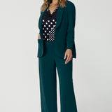 Showing a good work blazer for over 50s women, a size 12 woman wears a longline, green blazer with wide leg green work pants and a navy and white spot print top. These stretchy work jacket is smart for work and corporate wear. Made in Australia by Australian and New Zealand women's clothing brand, Leina & Fleur, shop work blazers in plus size to petite height in their online boutique.