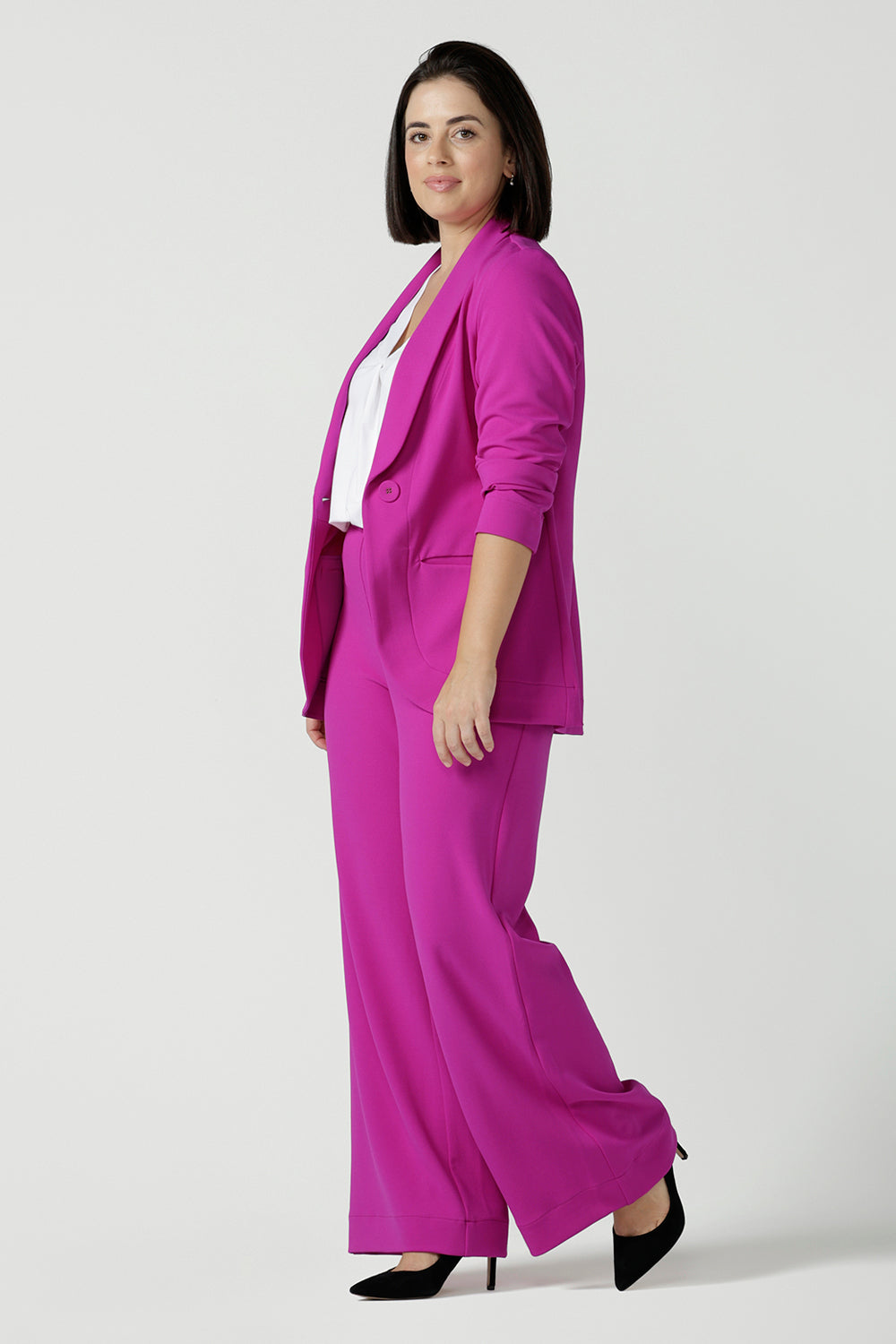 Size 10 woman wears a fuchsia Merit Blazer in scuba crepe. Tailored design with self covered buttons, shawl collar and curved hem and pockets. Size inclusive for stylish corporate workwear for women. Size 8 - 24.