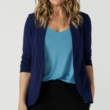 A size 10 woman wears the Merit Blazer in Sailor Scuba crepe fabric. A softly tailored blazer with lapel collar and full length sleeves. Button front and pockets. Made in Australia for women size 8-24. Styled back with a Fernanda Dress in Black wit a v-neck. Styled back with a Eddy Cami in Mineral. 