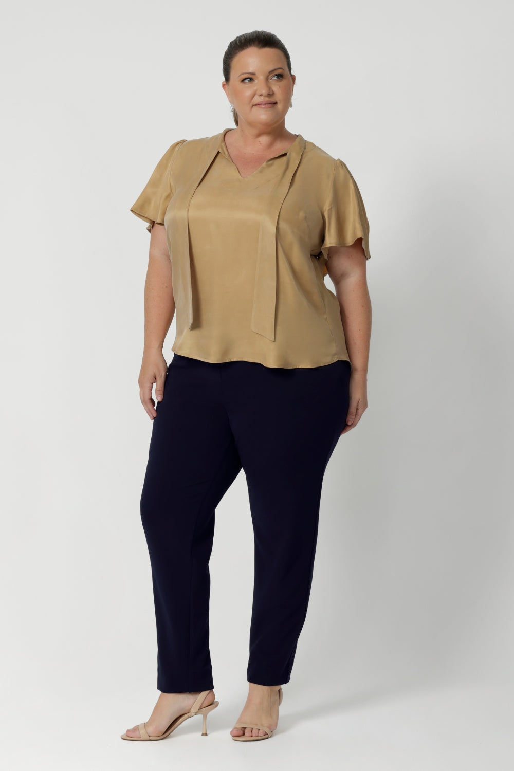 Plus size woman wears semi-fitted design of the Michael Shirt. Tailored yet relaxed feel, this blouse has the right amount of structure without feeling too tight. The V-neckline with delicate neckties adds a touch of feminine elegance, while allowing you to adjust the fit for added comfort and style. The short flutter sleeves, with slits on the shoulders, add a modern, airy feel to the blouse, perfect for this time of year. Available in sizes 8-24.