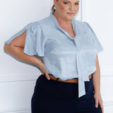 Image of plus size model wearing suede touch blouse styled with wide leg navy pants, brown belt and silver accessories. The V-neckline with adjustable neckties allows for a customisable fit, adding a feminine touch to the look. Short flutter sleeves with slits at the shoulders create a breezy, modern feel, bringing a subtle hint of playful elegance to the piece.