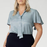 Image of plus size model wearing suede touch blouse styled with navy work skirt and silver accessories. The V-neckline with adjustable neckties allows for a customisable fit, adding a feminine touch to the look. Short flutter sleeves with slits at the shoulders create a breezy, modern feel, bringing a subtle hint of playful elegance to the piece.