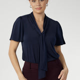 Close up shot of 30 year old woman wears navy work shirt with neck ties that can be styled in many different ways. This work shirt is perfect for a corporate look and pairs well with most Leina + Fleur core pieces. Easy care fabric is dry touch jersey. Garment is Australian made for women in Australia and New Zealand. Leina + Fleur is a women’s retail brand that proudly stocks sizes from 8-24. Shop this navy top in petite and plus sizes.