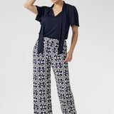 Full body front view of 30 year old woman wearing a navy top tucked into printed pants. This short sleeved shirt has neck ties that can be styled in a bow or left untied. Easy care dry touch jersey fabric was used to create this garment.