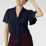 Woman wearing a navy blue top with neck ties detail and wine straight leg pants. Close up shot of 30 year old woman wears navy work shirt with neck ties that can be styled in many different ways. This work shirt is perfect for a corporate look and pairs well with most Leina + Fleur core pieces. 