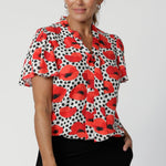 Povot print features vibrant poppies in red, tangerine, and black against a white base, adding a pop of colour and sophistication to a timeless design. Size 12 woman wears her printed workwear top with black pants. 