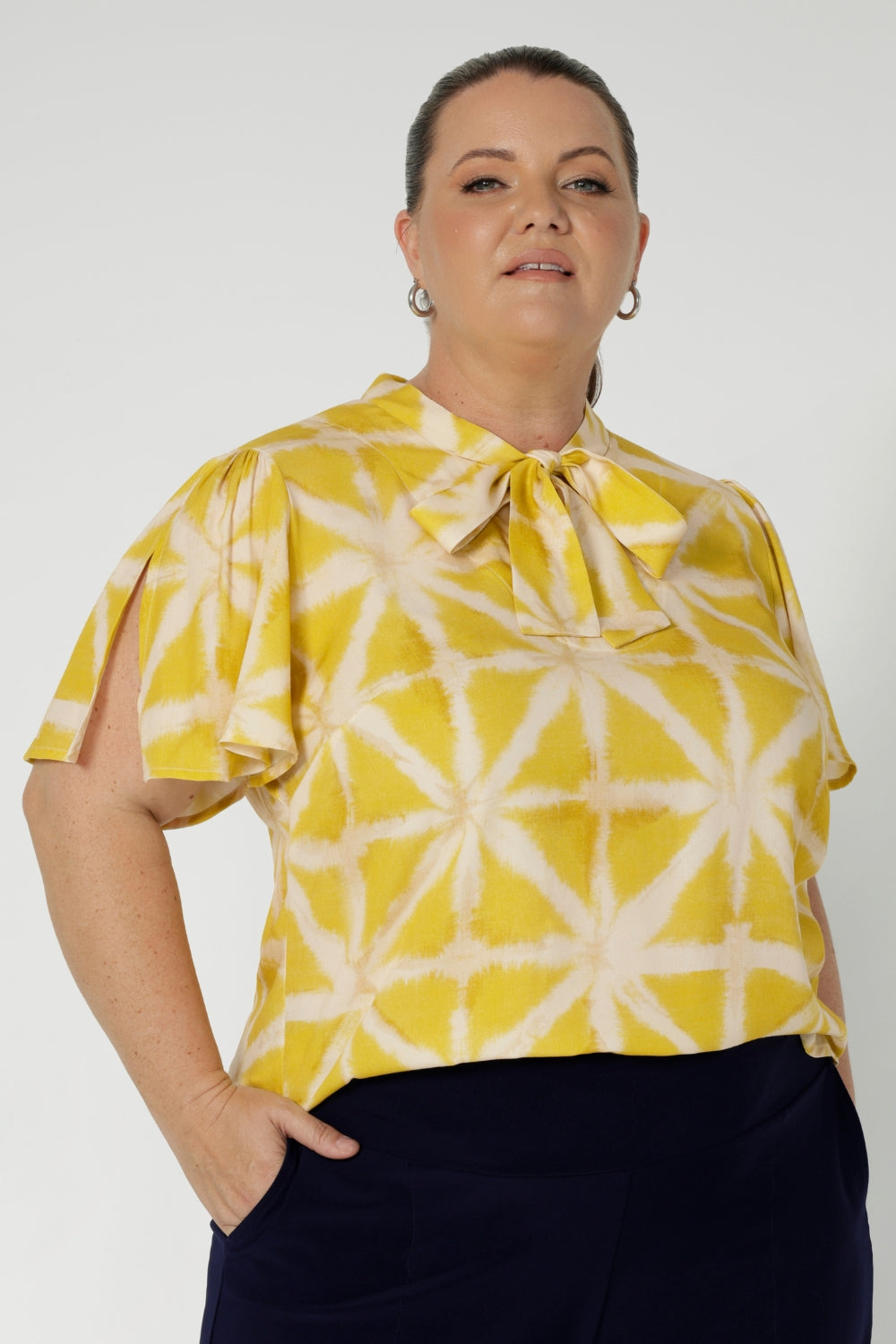 Plus sized model wears yellow coloured blouse featuring a V-neckline with delicate neckties and short flutter sleeves with slits on the shoulder, offering a breezy, relaxed feel perfect for warmer days. 