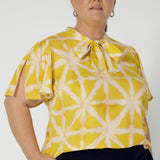 Plus sized model wears yellow coloured blouse featuring a V-neckline with delicate neckties and short flutter sleeves with slits on the shoulder, offering a breezy, relaxed feel perfect for warmer days. 