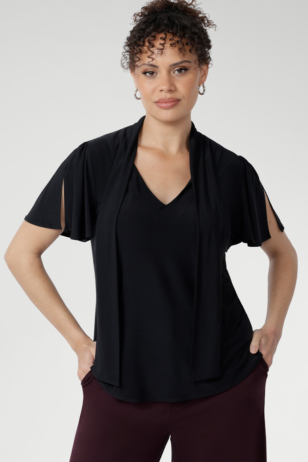 The Michael Top in Black is a timeless wardrobe staple designed for versatile styling. It features a soft tie neckline that can be worn loose, draped, or fastened high for a polished finish, along with short sleeves subtly gathered at the shoulder and finished with a split for elegant movement and coverage.