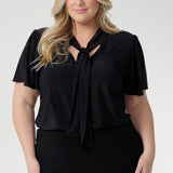 Designed for versatility, it features a soft tie neckline that can be worn loose and relaxed, gently draped, or fastened high for a more polished, elevated look. Short sleeves are subtly gathered at the shoulder and finished with a refined split, offering elegant movement and light coverage without compromising comfort.