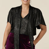 A petite height woman wears a good top for women's evening and event wear. In shimmering bronze foil print, this V-neck top with short flutter sleeves is made in Australia by women's occasionwear brand, Leina and Fleur.