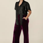 A petite height woman wears a good top for women's evening and event wear. In shimmering bronze foil print, this V-neck top with short flutter sleeves is worn with wide leg velvet evening trousers. Both evening top and cocktail pants are made in Australia by women's occasionwear brand, Leina and Fleur.