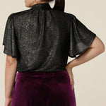 Back view of a petite height woman wearing a good top for women's evening and event wear. In shimmering bronze foil print, this V-neck top with short flutter sleeves is made in Australia by women's occasionwear brand, Leina and Fleur.