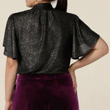 Back view of a petite height woman wearing a good top for women's evening and event wear. In shimmering bronze foil print, this V-neck top with short flutter sleeves is made in Australia by women's occasionwear brand, Leina and Fleur.