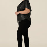 Side view of a plus size, size 18 woman wearing a good top for curvy women's evening and event wear. In shimmering bronze foil print, this V-neck top with short flutter sleeves is worn with slim leg black trousers for an elegant cocktail wear look. Made in Australia by women's occasionwear brand, Leina and Fleur.