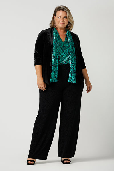 A curvy size 18 woman wears a good top for curvy women's evening and event wear. In  shimmering jade metallic print, this V-neck top with short flutter sleeves is worn with wide leg black trousers and a black velour jacket for an elegant cocktail wear look. Made in Australia by women's occasion-wear brand, Leina and Fleur.