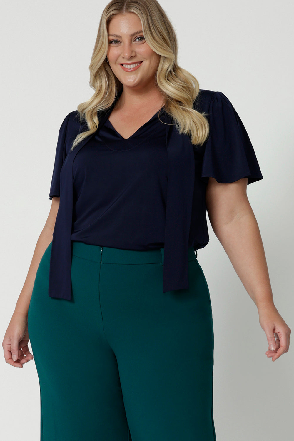 Close up shot of plus sized woman wearing navy blouse with neck ties. The workwear shirt is made of easy care dry touch jersey fabric. This work wear dress top is comfortable, easy and perfect for any corporate setting. Designed in Australia for Australian and New Zealand women. Leina & Fleur is a fashion brand that stock sizes from 8 to 24. Shop this tie up top in petite sizes and plus sizes.