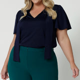 Close up shot of plus sized woman wearing navy blouse with neck ties. The workwear shirt is made of easy care dry touch jersey fabric. This work wear dress top is comfortable, easy and perfect for any corporate setting. Designed in Australia for Australian and New Zealand women. Leina & Fleur is a fashion brand that stock sizes from 8 to 24. Shop this tie up top in petite sizes and plus sizes.