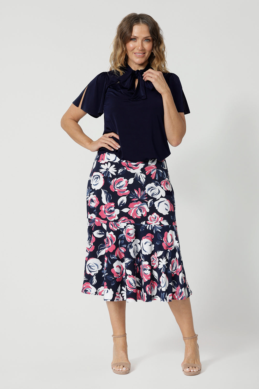 Full body front view of 40 year old woman wearing a printed floral skirt. This 3/4 length midi skirt is made from dry touch jersey fabric and has a colourful combination of white and pink roses on navy base. This breathable work wear skirt is comfortable and is considered a travel essential. This easy care fabric is made in Australia by Australian and New Zealand women. Leina and Fleur is a woman clothing brand that stocks sizes from 8 to 24. Shop this printed skirt in plus and petite sizes. 