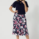 Full body front view of 40 year old woman wearing a printed floral skirt. This 3/4 length midi skirt is made from dry touch jersey fabric and has a colourful combination of white and pink roses on navy base. This breathable work wear skirt is comfortable and is considered a travel essential. This easy care fabric is made in Australia by Australian and New Zealand women. Leina and Fleur is a woman clothing brand that stocks sizes from 8 to 24. Shop this printed skirt in plus and petite sizes. 