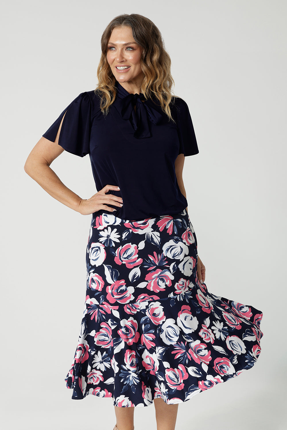 Close up front on image of 40y/o woman wearing a brightly printed midi skirt and navy top. Breathable material used is easy to care dry touch jersey. The floral printed skirt has colours of white, navy and pink. Lightweight skirt is Australian made by women for Australian and New Zealand women. Leina and Fleur is a woman's fashion brand that actively stocks plus and petite sizes ranging from 8 to 24. 