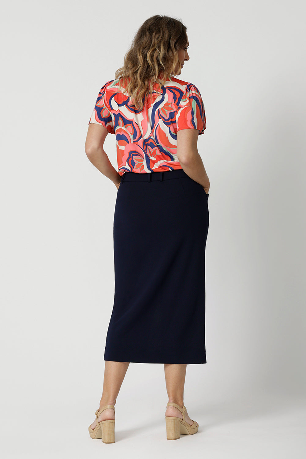 Full body back view of woman wearing midi length skirt. Navy skirt is made to stretch comfortably around the body. Designed with high quality, easy care scube crepe fabric.  Workwear skirt has an above the knee length split. This comfortable work wear skirt was made for any office or corporate setting. Australia made clothing by woman for woman. Made in Australian by Australian and New Zealand women. Leina & Fleur is a woman’s clothing brand that have sizes ranging from 8 to 24. Shop appropriate work skirt.