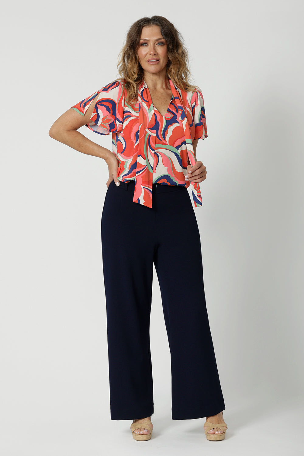 Showing a good summer top for work, the Michael Top has flutter sleeve and a neck tie detail. Showing stylish tops for over 50s women, this jersey top is worn by a size 12, over 50 woman. Worn with classic, navy wide leg pants, Wear for work or cruise wear . This top is made in Australia by Australian and New Zealand clothes company, Leina & Fleur. 