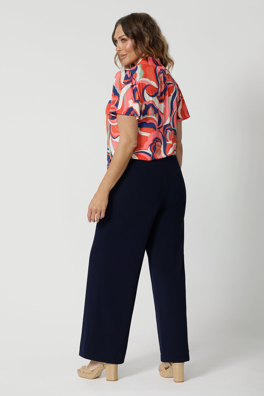 Back view of a good summer top for work, the Michael Top has flutter sleeve and a neck tie detail. Showing stylish tops for over 50s women, this jersey top is worn by a size 12, over 50 woman. Worn with classic, navy wide leg pants, this top wears for work or cruise wear. This flutter sleeve top is made in Australia by Australian and New Zealand clothes company, Leina & Fleur. 