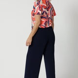 Back view of a good summer top for work, the Michael Top has flutter sleeve and a neck tie detail. Showing stylish tops for over 50s women, this jersey top is worn by a size 12, over 50 woman. Worn with classic, navy wide leg pants, this top wears for work or cruise wear. This flutter sleeve top is made in Australia by Australian and New Zealand clothes company, Leina & Fleur. 