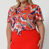 Showing a good summer top for plus size women, the Michael top has flutter sleeves and stylish necktie.  Worn by a size 18, plus size women, this curve top is worn with tangerine, orange tapered leg pants. Wear this comfortable top for work or cruise wear . This short sleeve top for women is made in Australia by Australian and New Zealand clothes company, Leina & Fleur. 