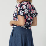 Close up side view of forty year old woman wearing a short sleeve top. Top has neck ties and is made of dry touch jersey material. Blouse has a floral design with roses in the colours pink, white and navy. Easy care fabric is used to craft Australian made clothing for Australian and New Zealand women. Woman’s clothing retailer Leina and Fleur stock sizes petite and plus sizes from 8 to 24.