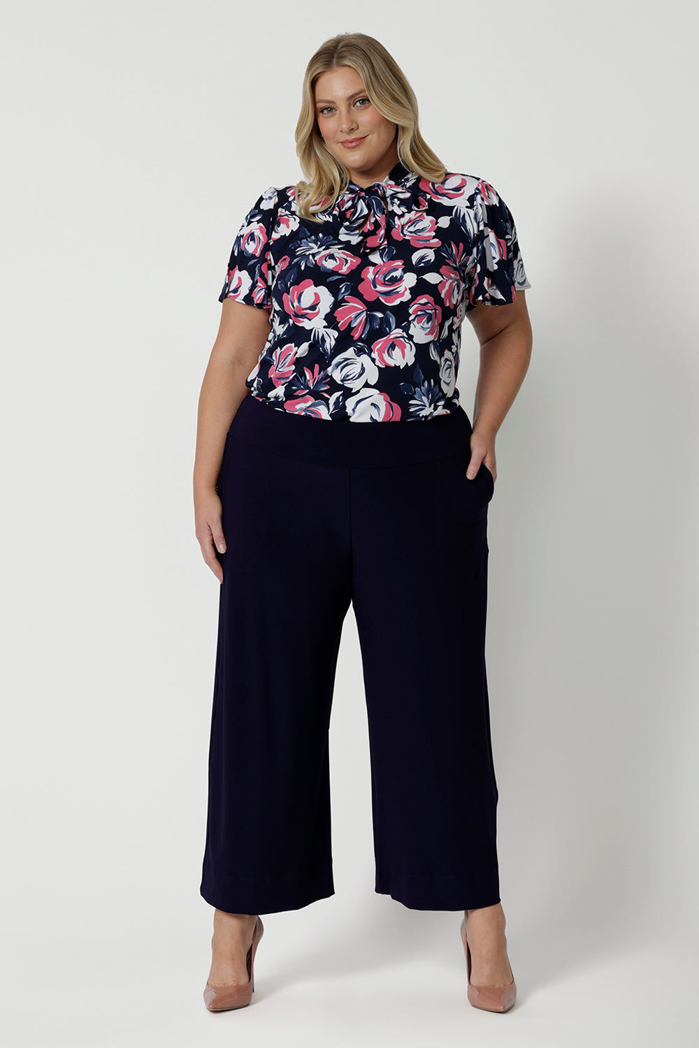 Full body front view of plus sized female model, wearing a floral short sleeved top made of high quality dry touch jersey fabric. Shirt is navy coloured with pink and white roses. This easy care shirt is breathable and Australian made. Made for woman in Australia and New Zealand. Leina & Fleur is an online woman’s clothing brand that stocks petite & plus sizes ranging from 8 to 24.