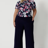 Full body front view of plus sized female model, wearing a floral short sleeved top made of high quality dry touch jersey fabric. Shirt is navy coloured with pink and white roses. This easy care shirt is breathable and Australian made. Made for woman in Australia and New Zealand. Leina & Fleur is an online woman’s clothing brand that stocks petite & plus sizes ranging from 8 to 24.